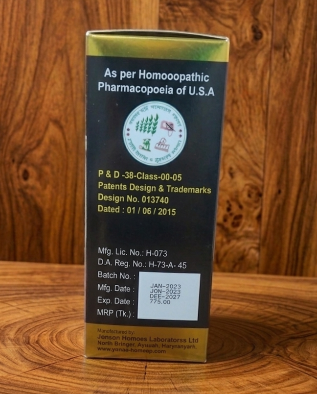 A tall dark box with gold cap standing on wood, labeled 'As per Homoeopathic Pharmacopoeia of U.S.A' and small circular logo. It lists patent design numbers and manufacture details on a yellow panel at the bottom.